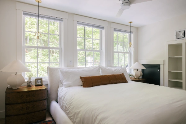 King bedroom framed by three large windows, pendant lighting, sculptural nightstands, and forest views beyond the glass